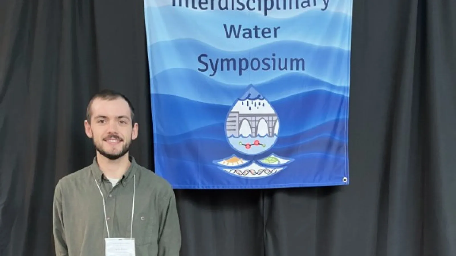 Seth Drake posing next to a flag for the Interdisciplinary Water Symposium