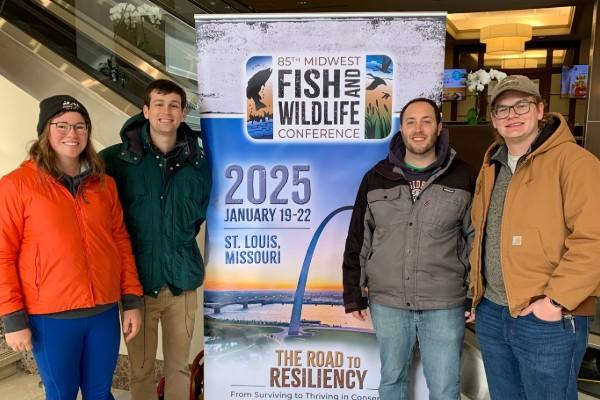 Shaley Valentine, Andrew Foley, Steven Gratz, and Jacob Bentley in front of a sign for the Midwest Conference