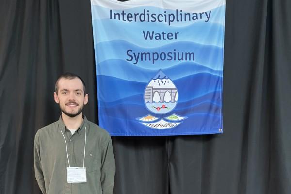 Seth Drake posing next to a flag for the Interdisciplinary Water Symposium